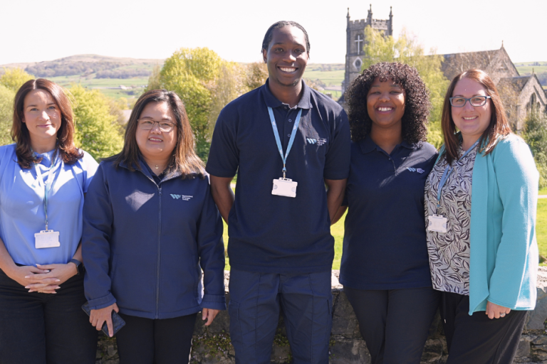 Five Community Support Workers standing together outdoors, wearing staff identification badges, with a rural landscape and church building in the background